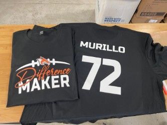 Black T-shirts laid out on a table, one reading “Difference Maker” and the other “MURILLO 72”