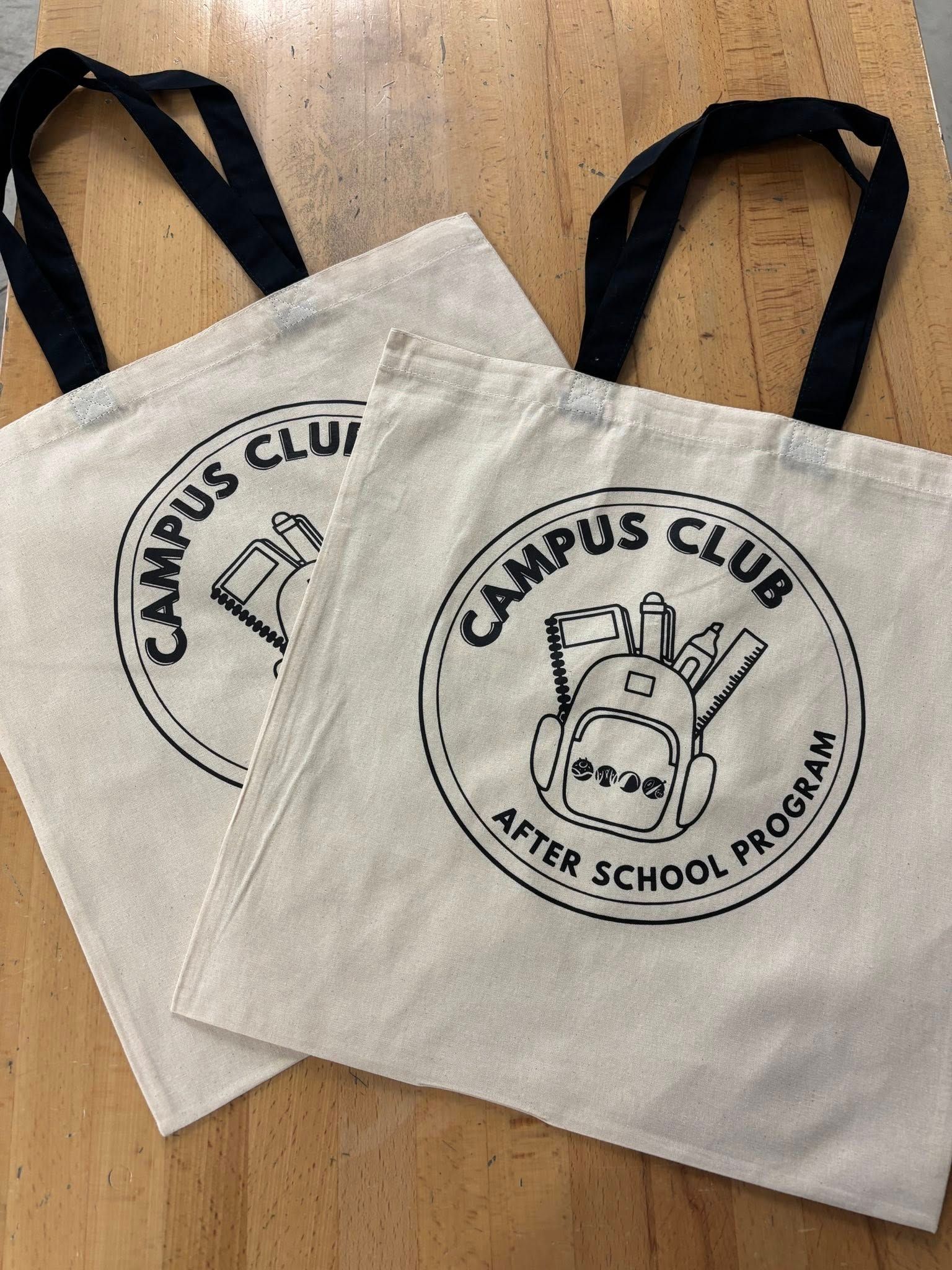 Two cream canvas tote bags with black handles and campus club after-school program logo on a wooden table
