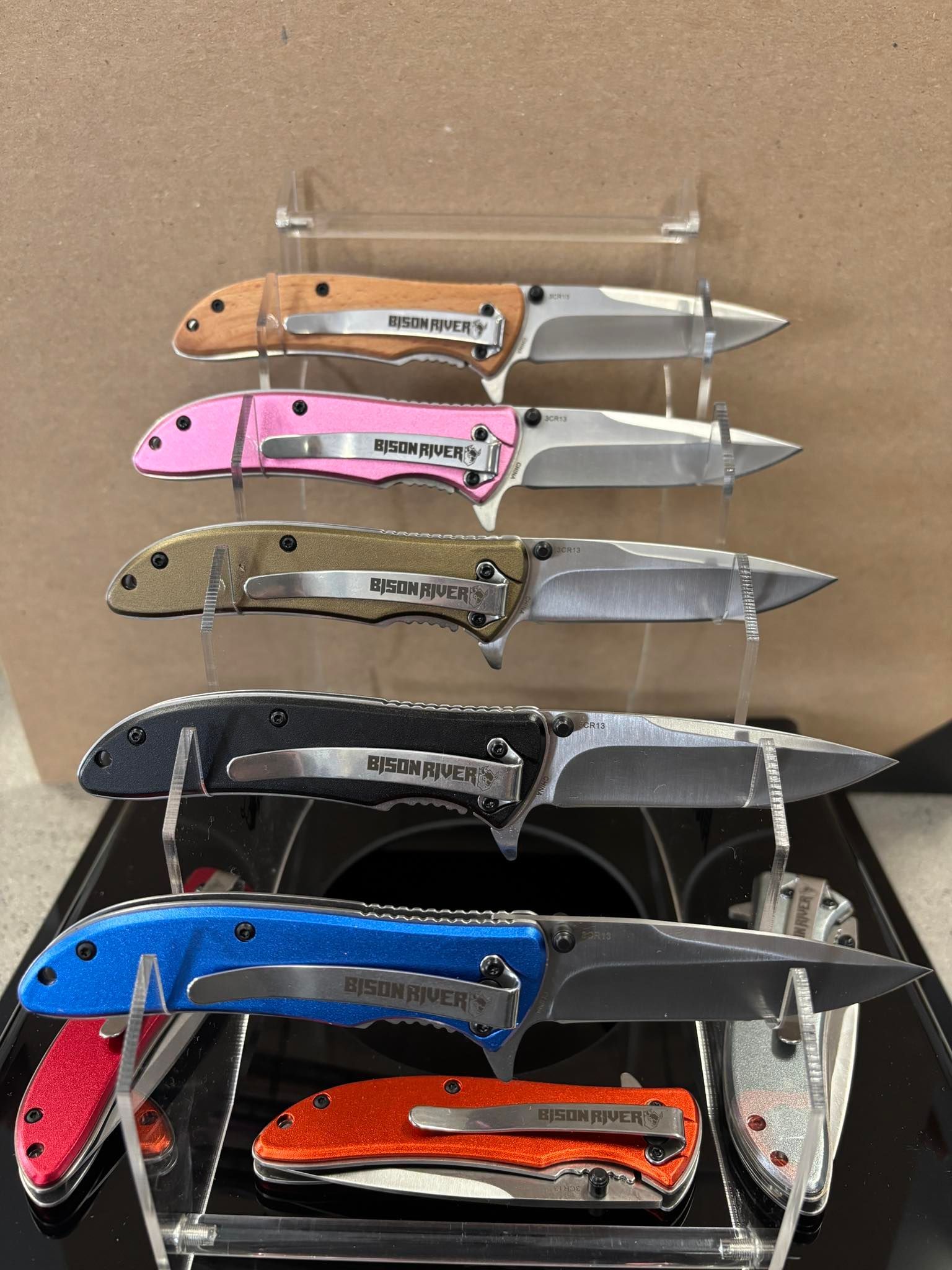 Stacked folding knives on display, with tan, pink, gold, black, blue, and orange handles.