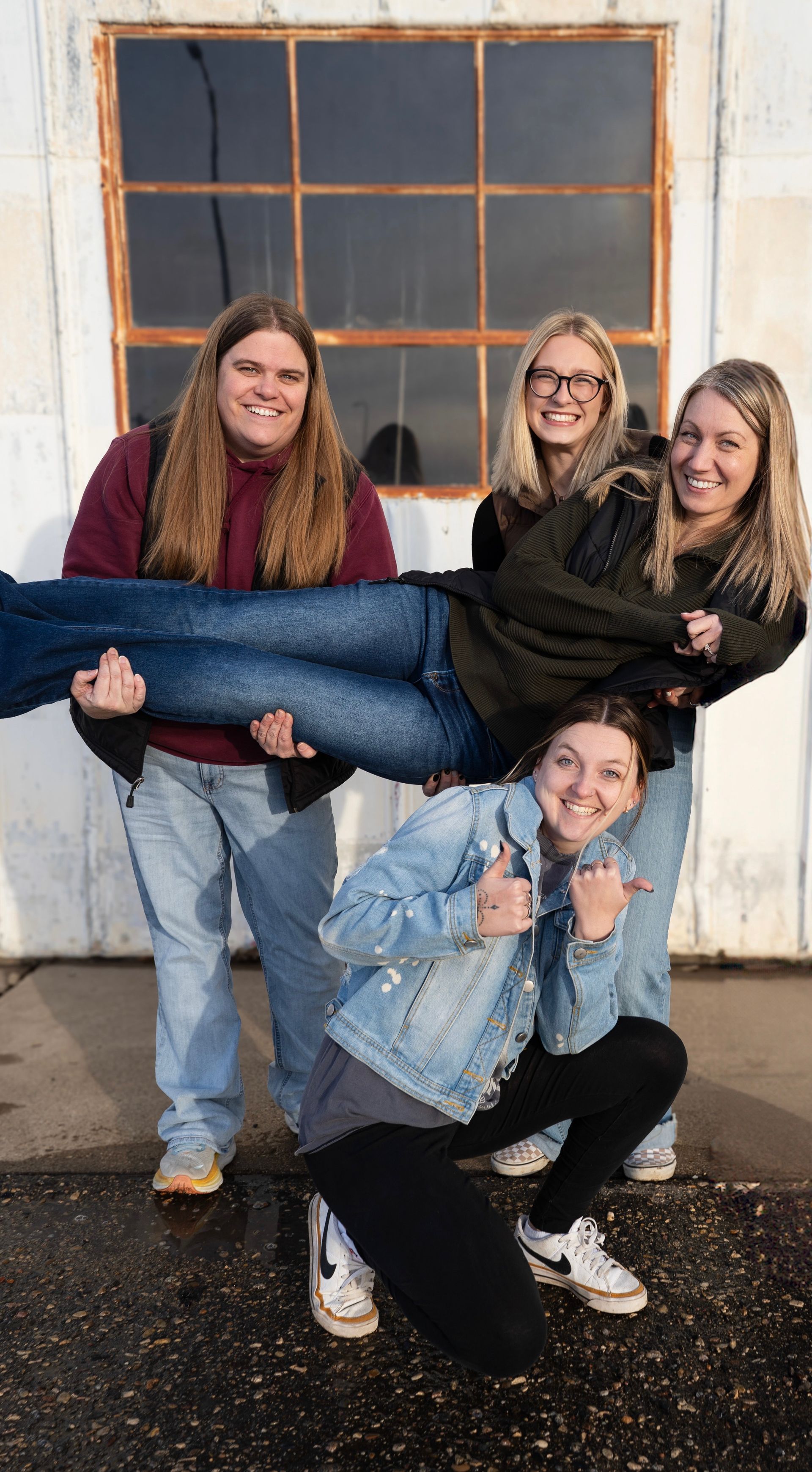 Three people posing indoors, with one being held horizontally and all smiling in front of a window.