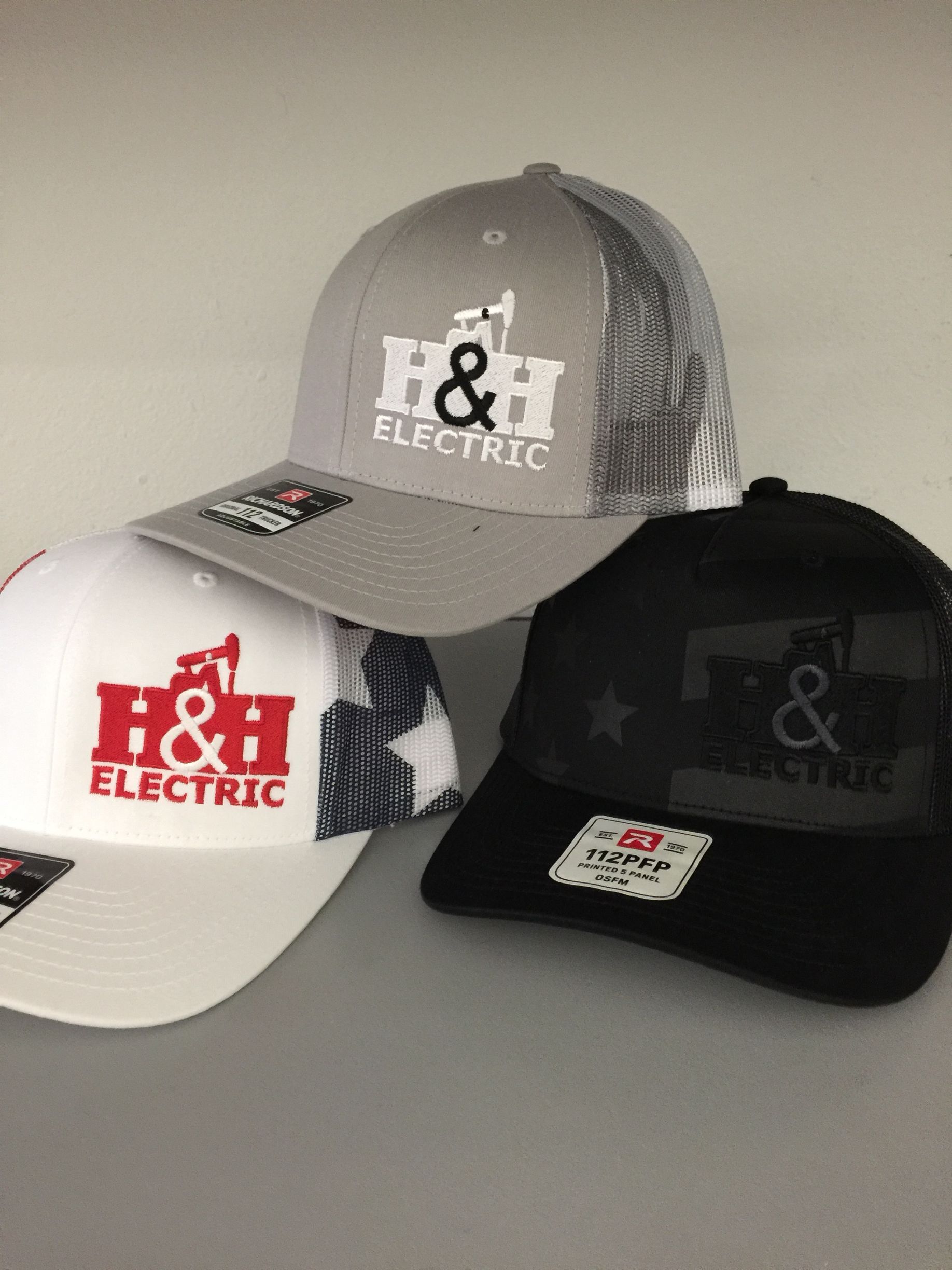 Three H&H Electric baseball caps in white, gray, and black, stacked on a light surface.