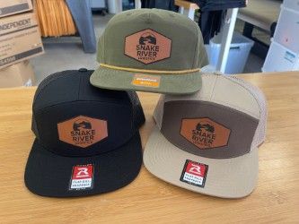Three trucker hats on a table: black, olive green, and tan with leather patch logos.