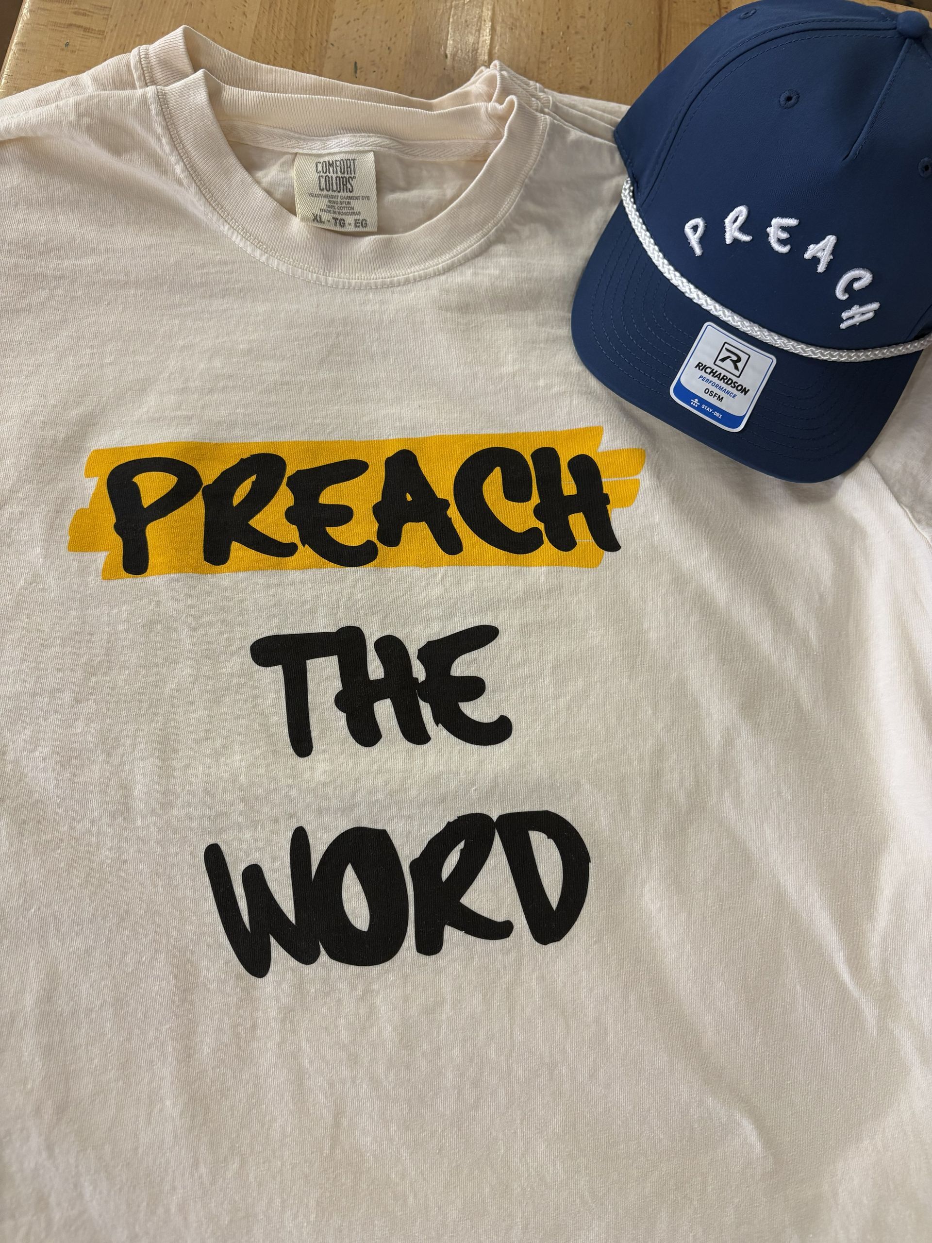 White T-shirt with “PREACH THE WORD” in black and yellow, next to a navy cap with “PREACH” embroidered in white.