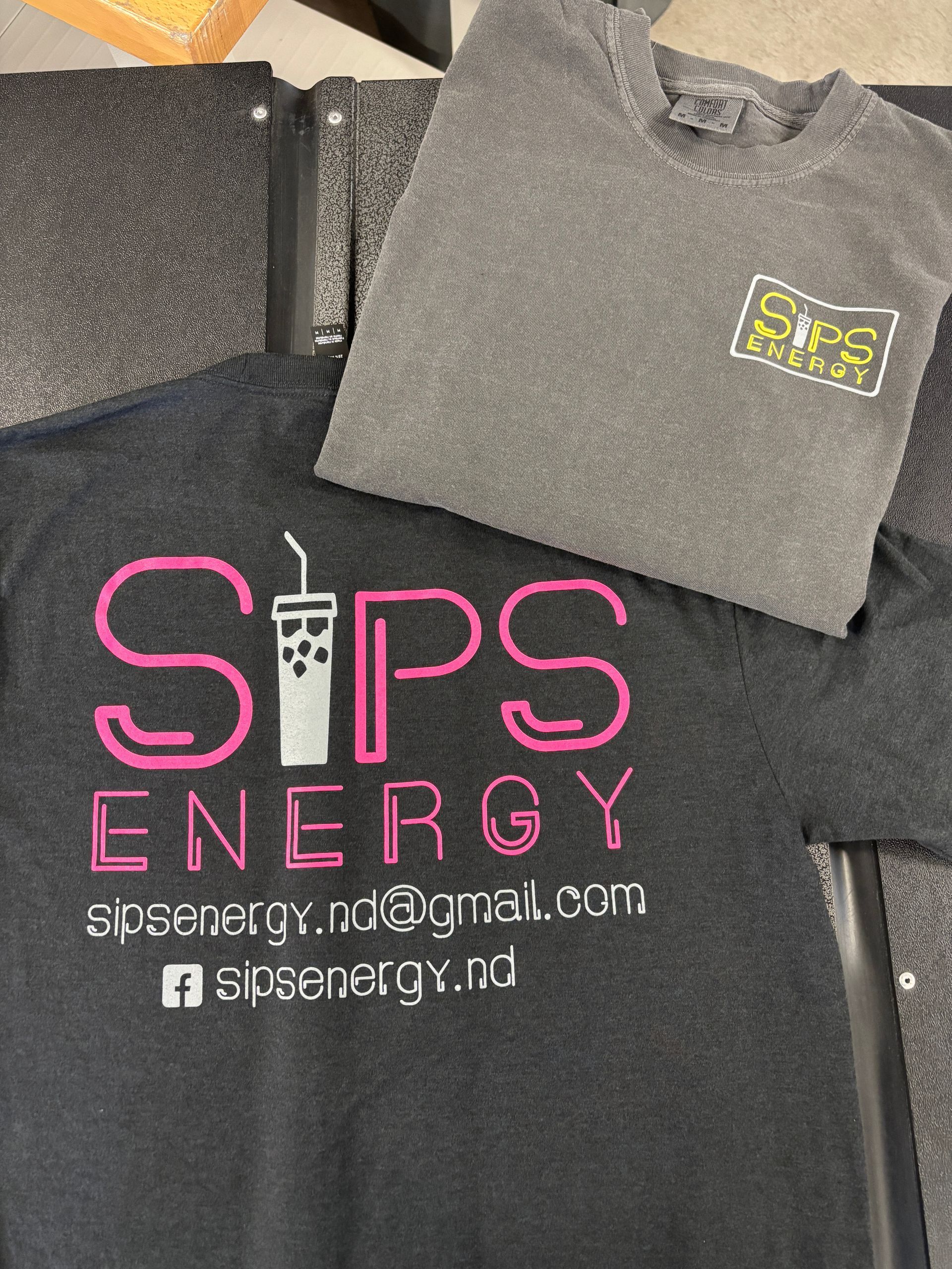 Black SITPS Energy T-shirt with pink logo and gray folded shirt on top on a textured surface