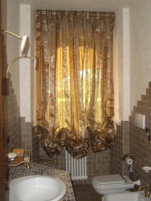 Bagno con finestra e tende dorate, pareti piastrellate marroni, water bianco e lavandino.