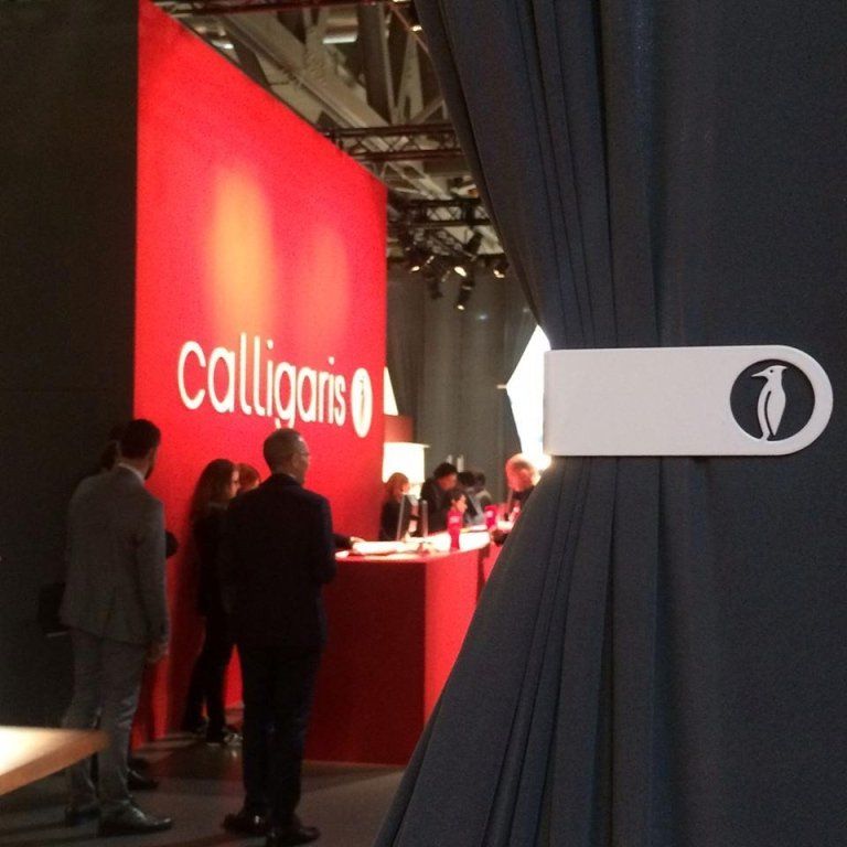 Persone presso uno stand rosso della Calligaris a una fiera, con una tenda con il logo di un pinguino.