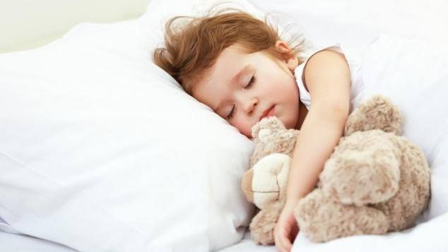 Un bambino dorme, coccolando un orsacchiotto, su una coperta bianca.