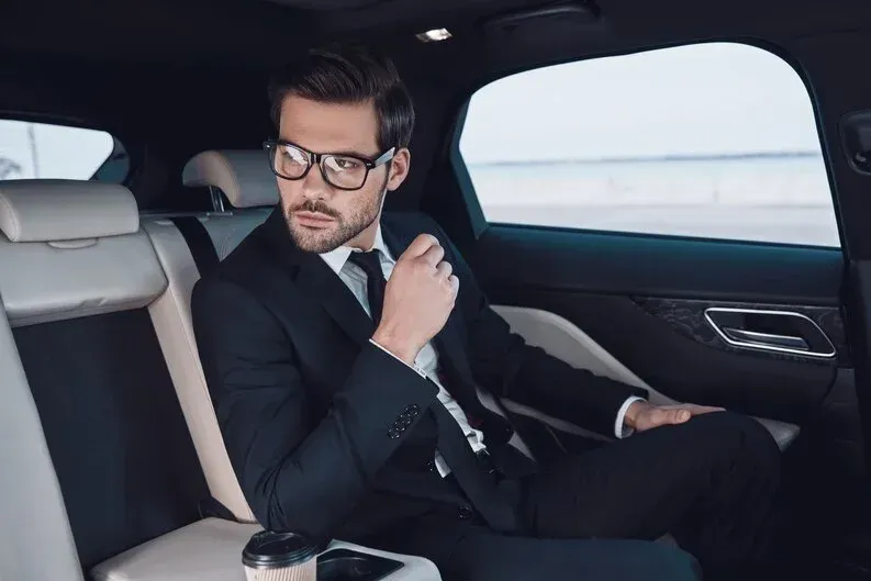 Man in a suit sits in the back seat of a car, looking out the window, holding his chin.