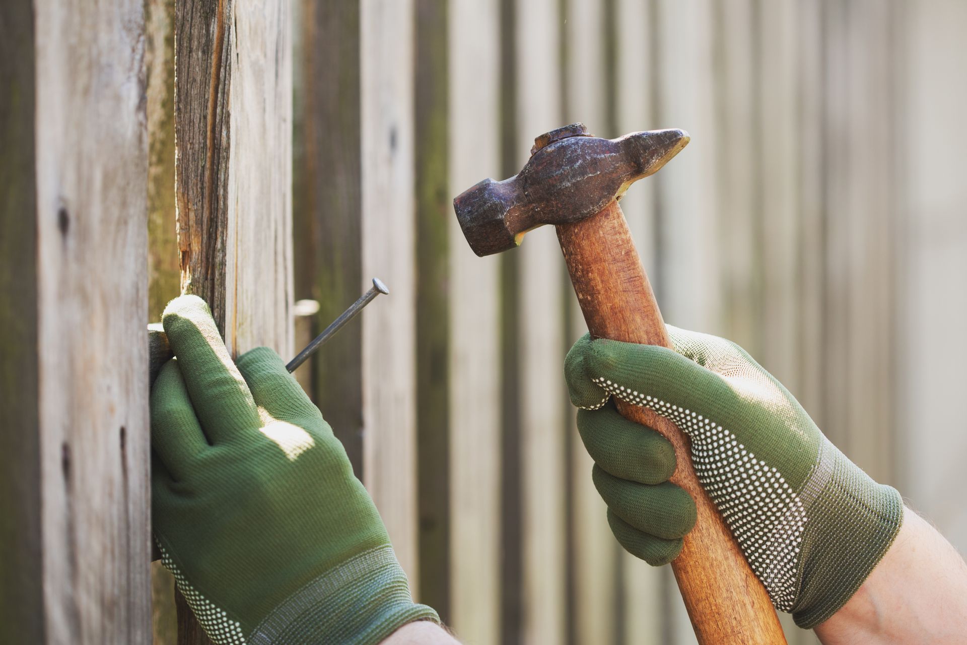 Hands in green gloves hammer a nail into a wooden fence panel.