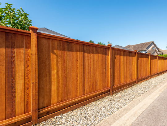Long wooden privacy fence along a gravel roadside under a clear blue sky