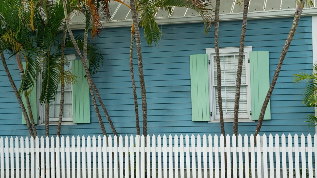 Blue house with mint shutters behind a white picket fence and palm trees in front