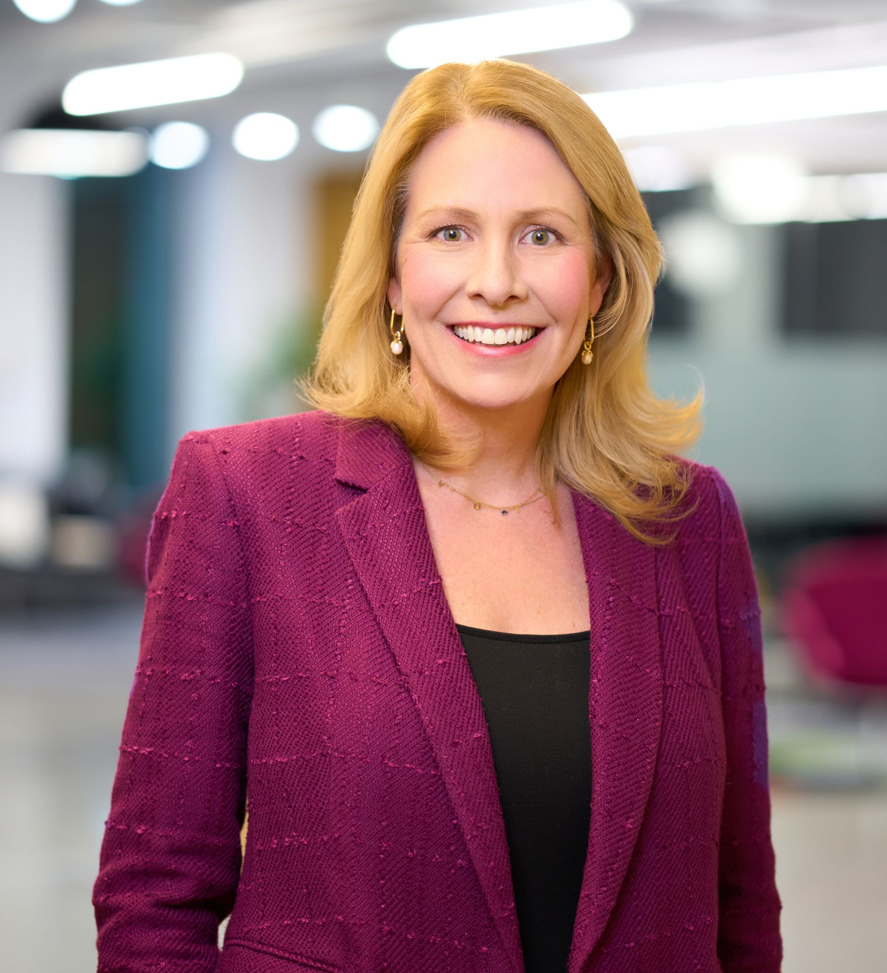 A blond haired woman wearing a burgundy blazer smiles in front of a blurred background. 