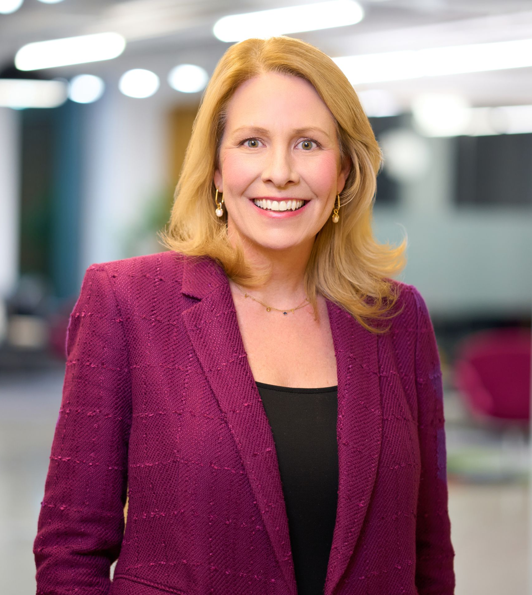 A woman wearing a burgundy blazer and a necklace smiles in front of a blurred office background.
