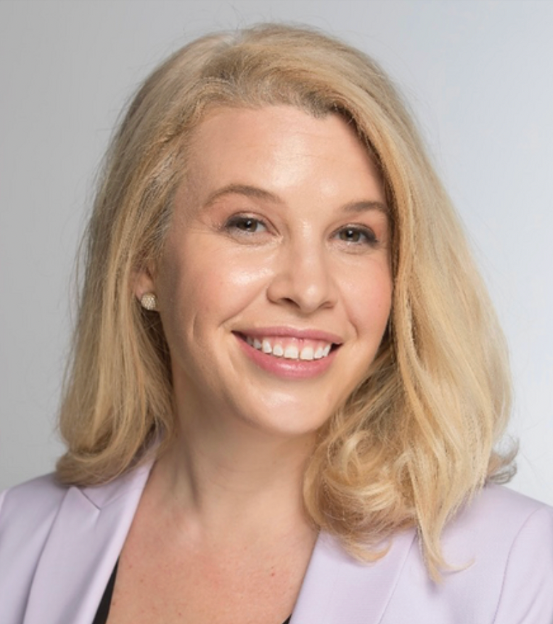 A woman with short blonde hair in a lilac blazer smiles in front of a gray background.