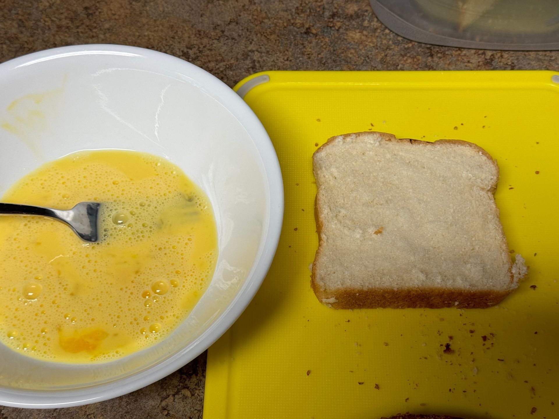 A bowl of eggs next to a slice of bread on a yellow cutting board