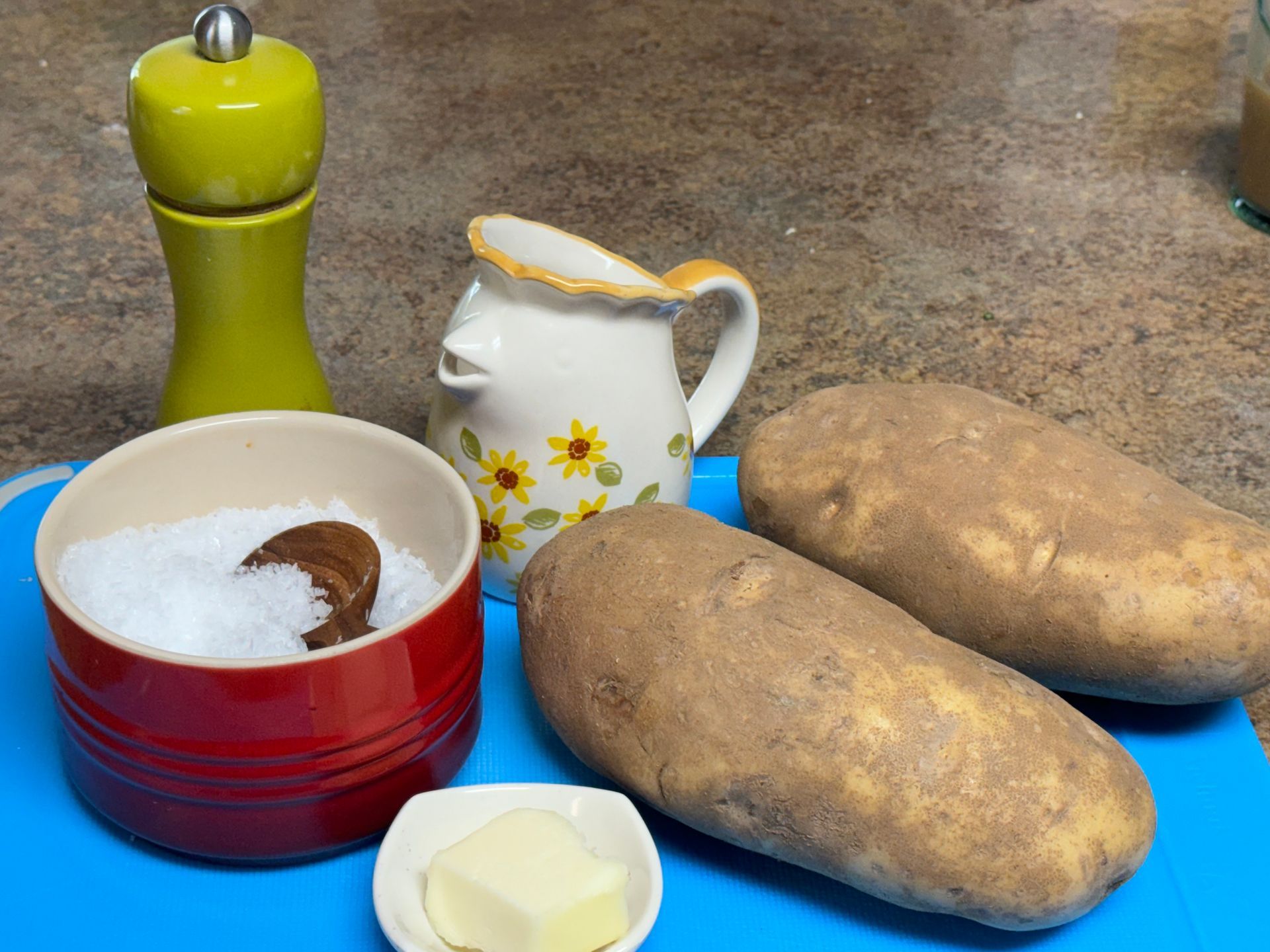 Potatoes , butter , salt and pepper are on a blue cutting board.