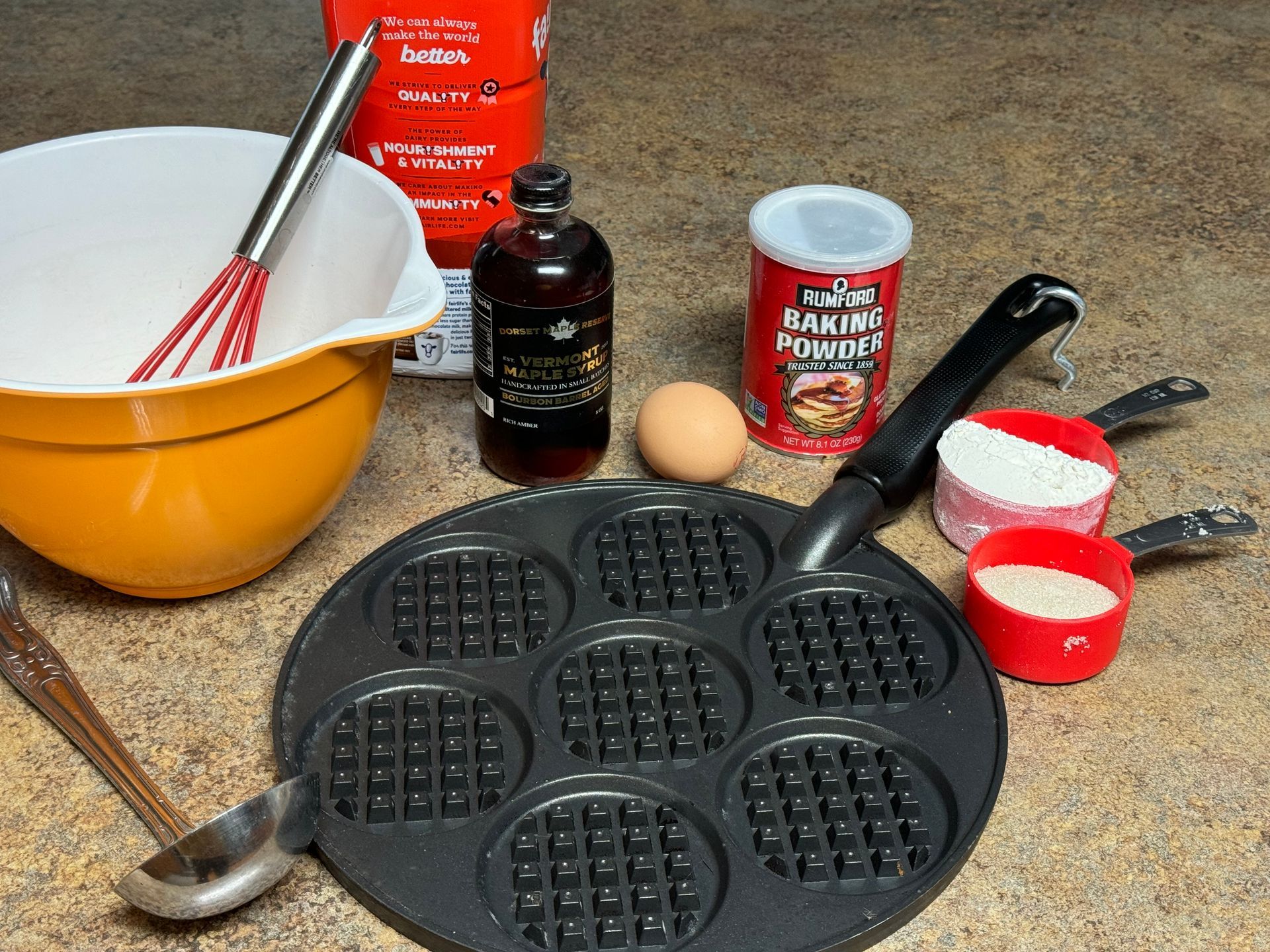 A waffle maker is surrounded by baking supplies including a bottle of baking powder