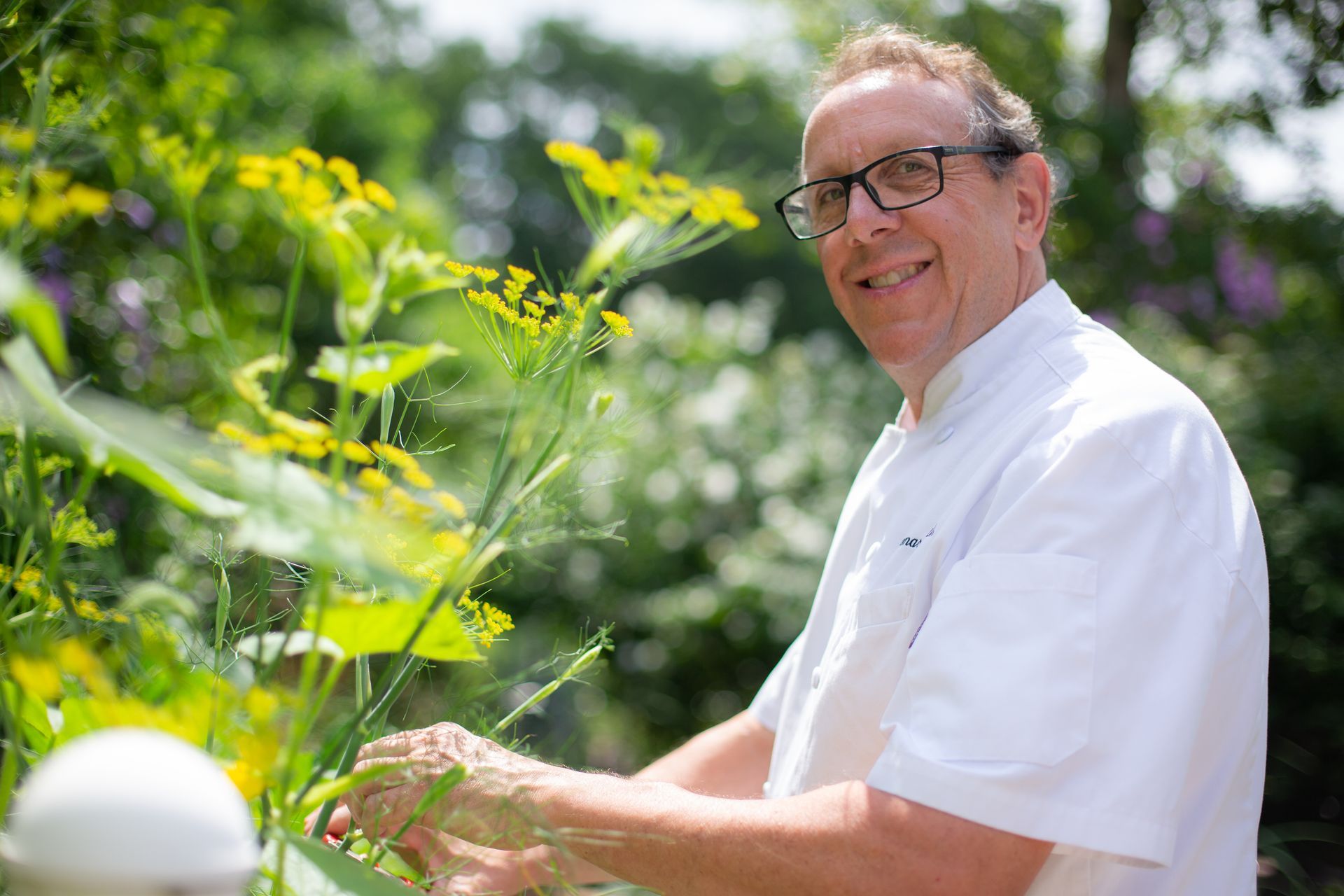 A man in a white shirt and glasses is standing in front of a bunch of flowers.