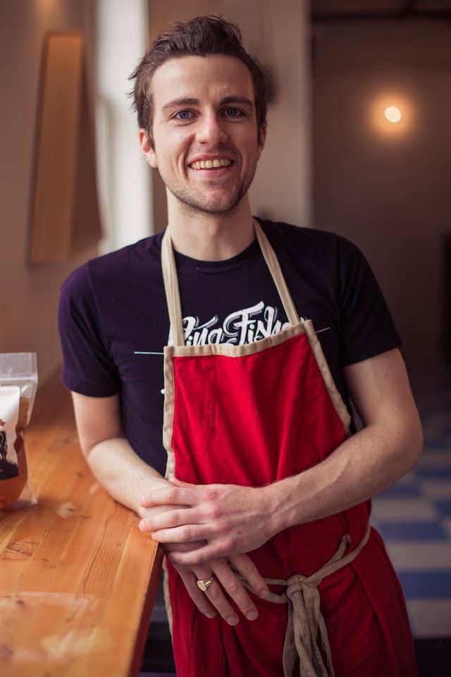 A man wearing a red apron and a black shirt is standing in front of a counter.