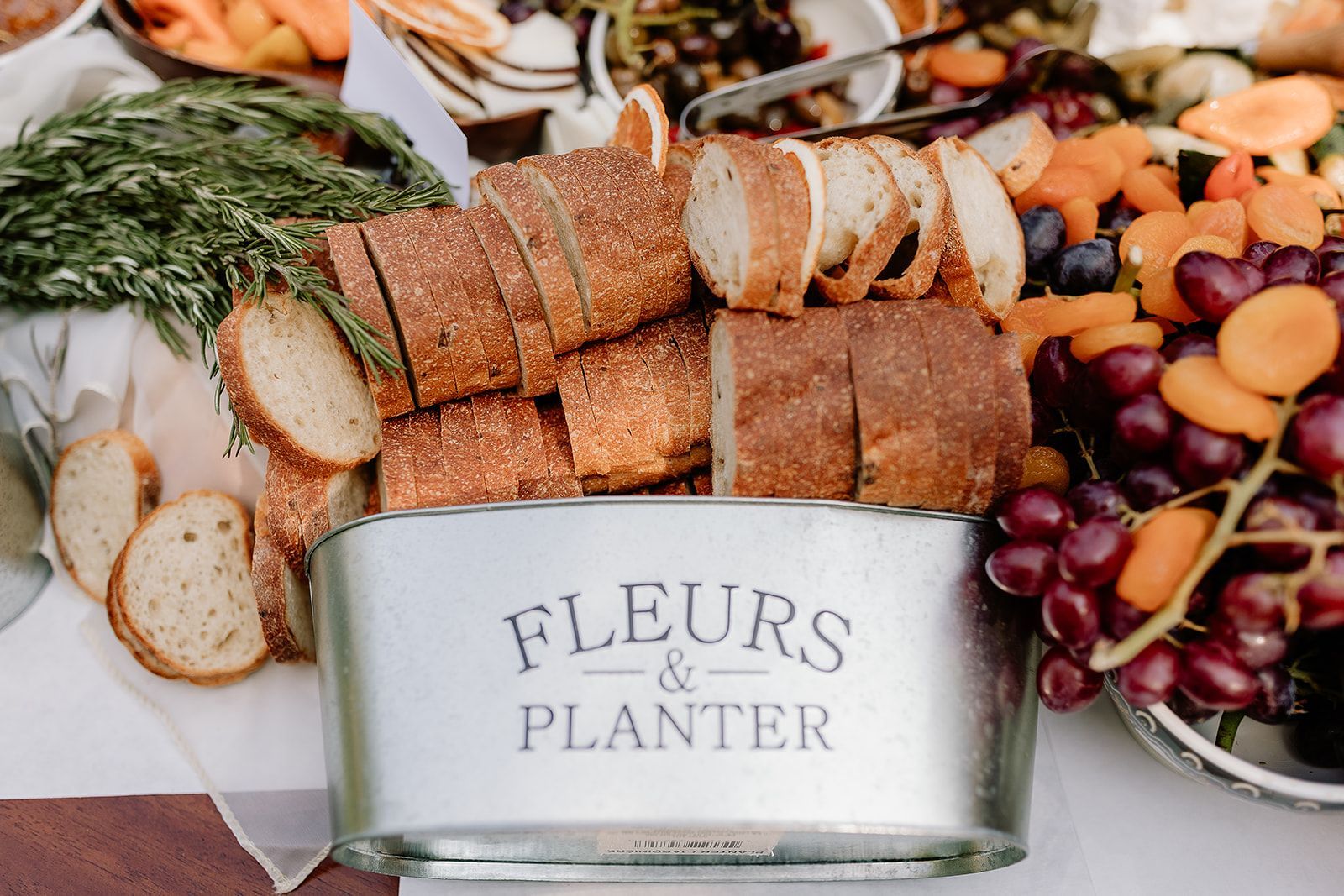 A tin labeled “Fleurs & Planter” with sliced bread, rosemary, and assorted fruits arranged around it.
