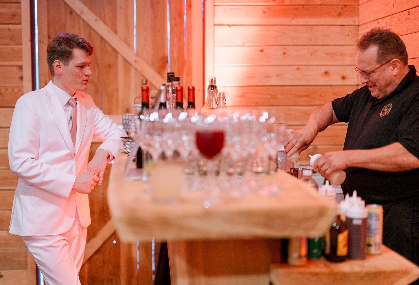 Two men at a bar counter with wine glasses, one in a white suit and one pouring a drink