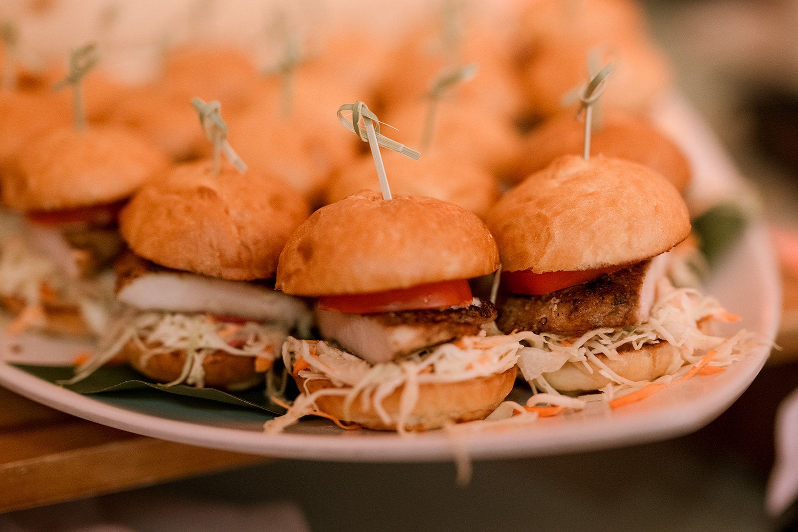 A white serving platter filled with multiple mini sliders topped with toothpicks, tomato slices, and shredded lettuce.