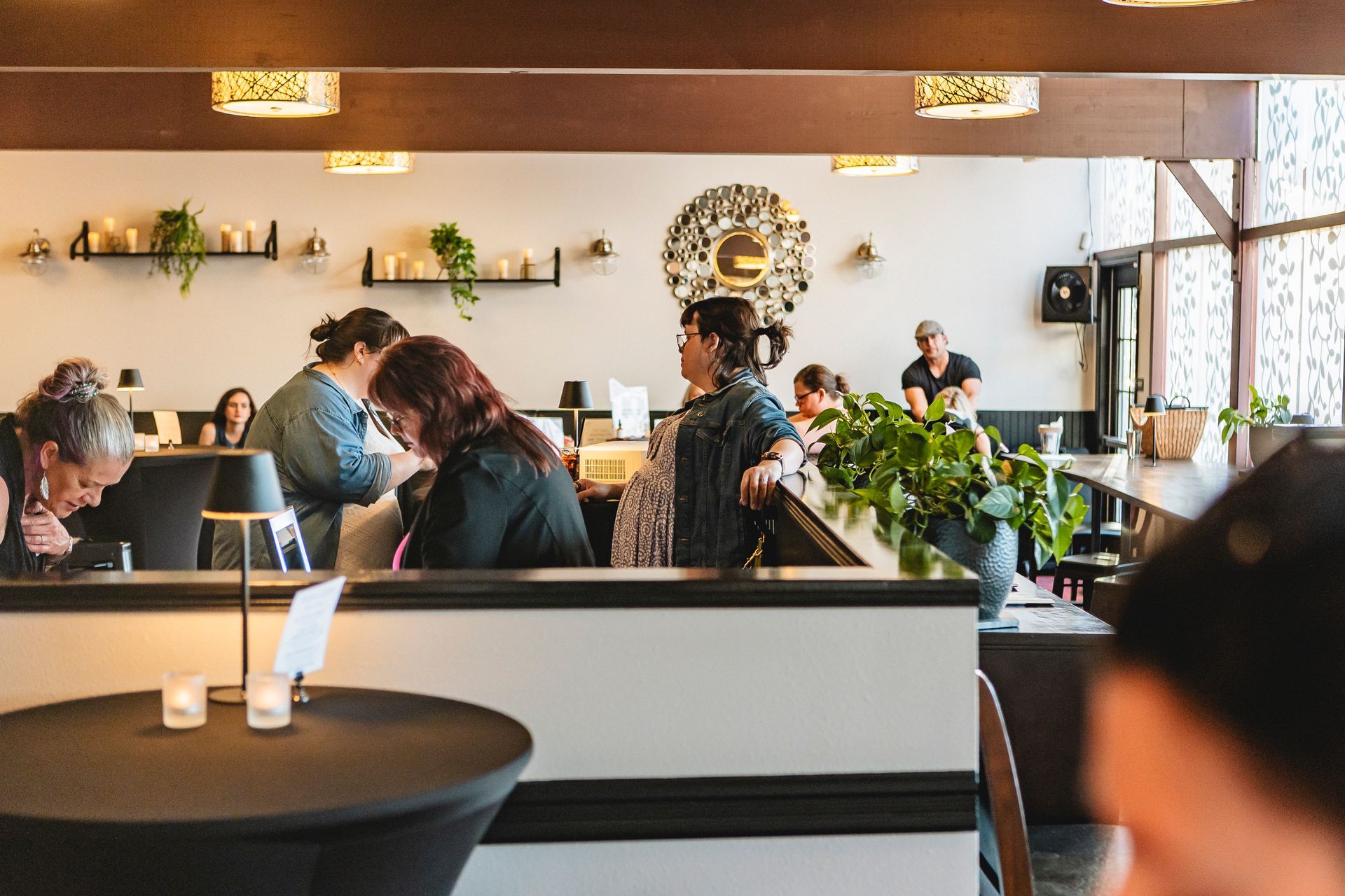 Busy café interior with people seated at tables, warm lighting, and potted plants near the counter.
