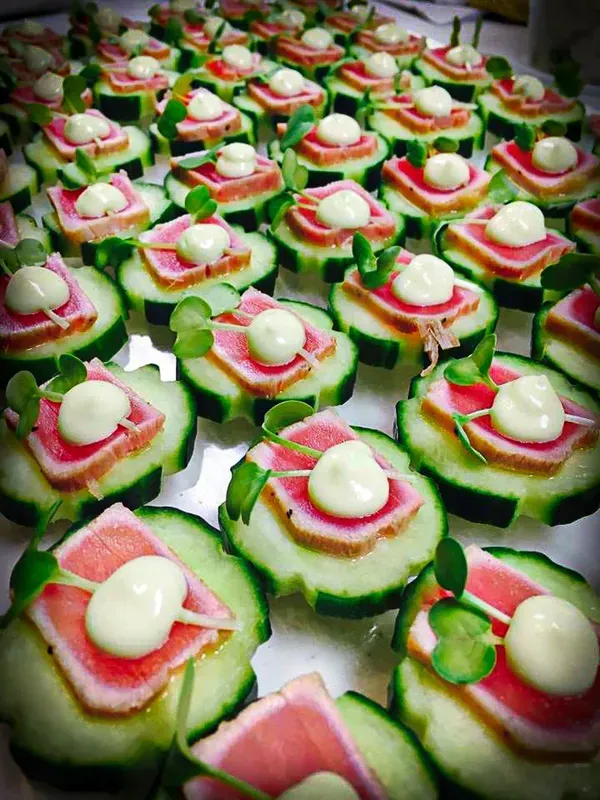 A large tray of seared tuna appetizers, each on a cucumber slice topped with a dollop of white sauce and microgreens.