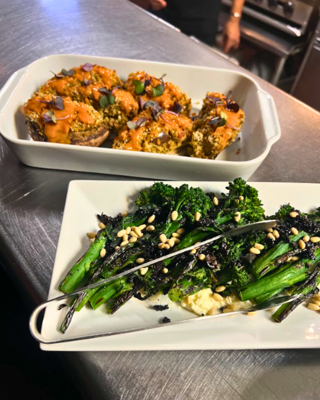 Two white dishes on a stainless steel surface, featuring stuffed mushrooms with sauce and sautéed broccolini with pine nuts.