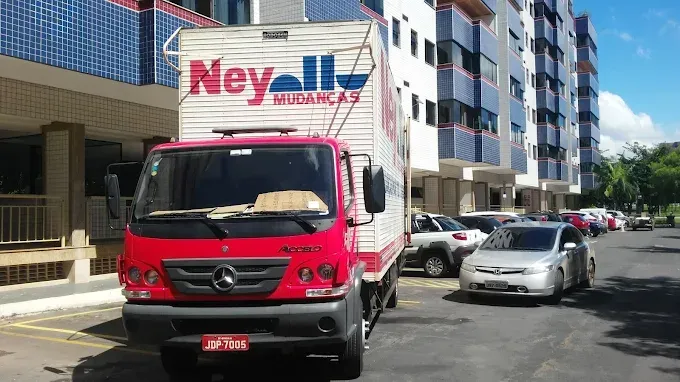 Caminhão de mudanças da Ney Mudanças vermelho estacionado na rua ao lado de um prédio e outros carros estacionados.