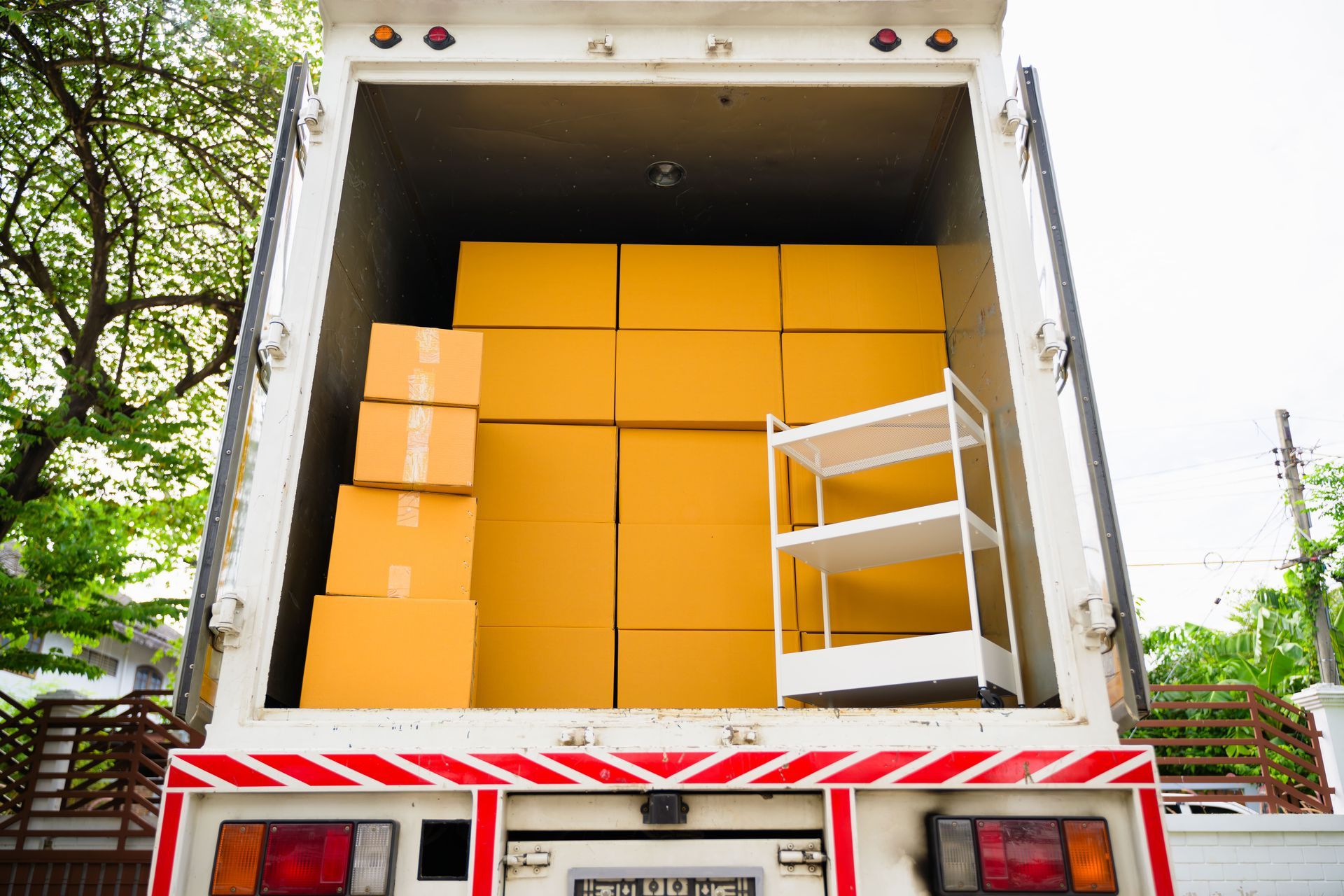 Carroceria aberta de um caminhão de entregas, repleta de caixas de papelão amarelas e uma estante de metal branco vazio.