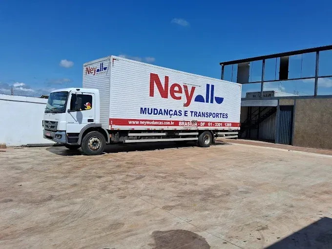 Caminhão de mudanças branco da Ney Mudanças estacionado num terreno de terra batida em frente a um prédio, sob um céu azul.