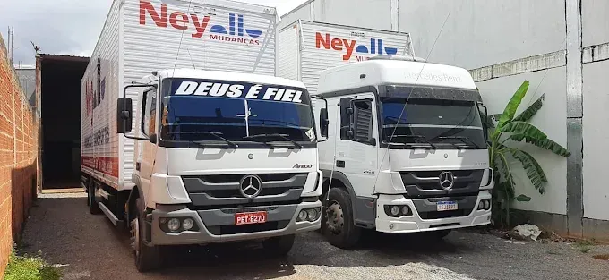 Dois caminhões Mercedes-Benz brancos da transportadora Ney Mudanças estacionados lado a lado sob uma área de cascalho.