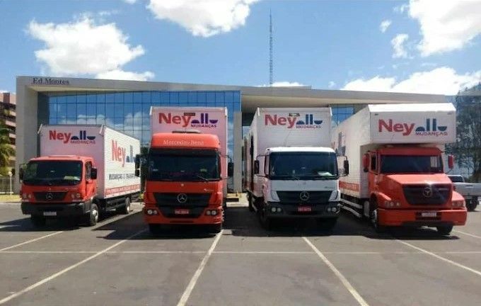 Quatro caminhões da Ney Mudanças estacionados em fila em frente a um moderno edifício com fachada de vidro, sob um céu azul.
