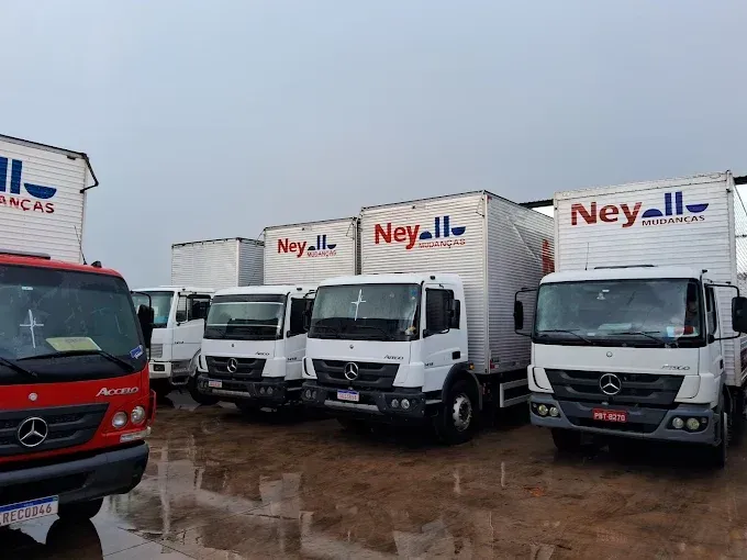 Caminhões Mercedes-Benz brancos estacionados e um caminhão vermelho em primeiro plano, todos  com logo da Ney Mudanças.