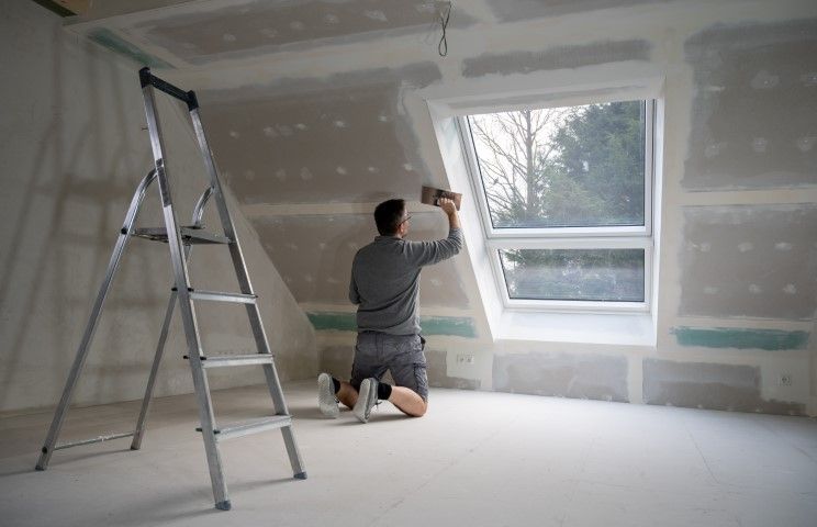 Person kneeling and painting a wall in a partially finished room beside a step ladder and window