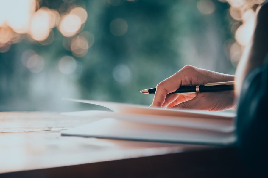 Hand holding a pencil above an open notebook on a desk, with soft blurred lights in the background