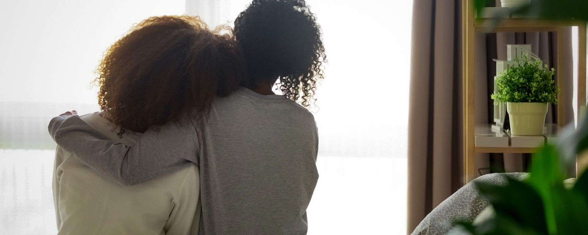 Two people sit with arms around each other by a bright window, with a potted plant nearby.