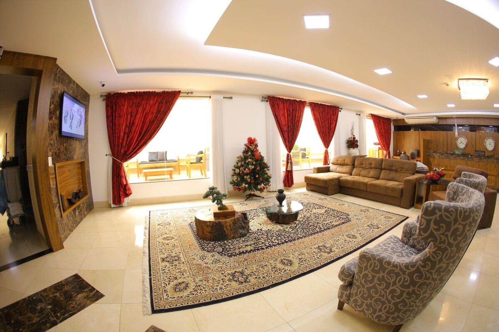 Sala de estar espaçosa com cortinas vermelhas, sofás bege, tapete estampado e iluminação aconchegante no teto.