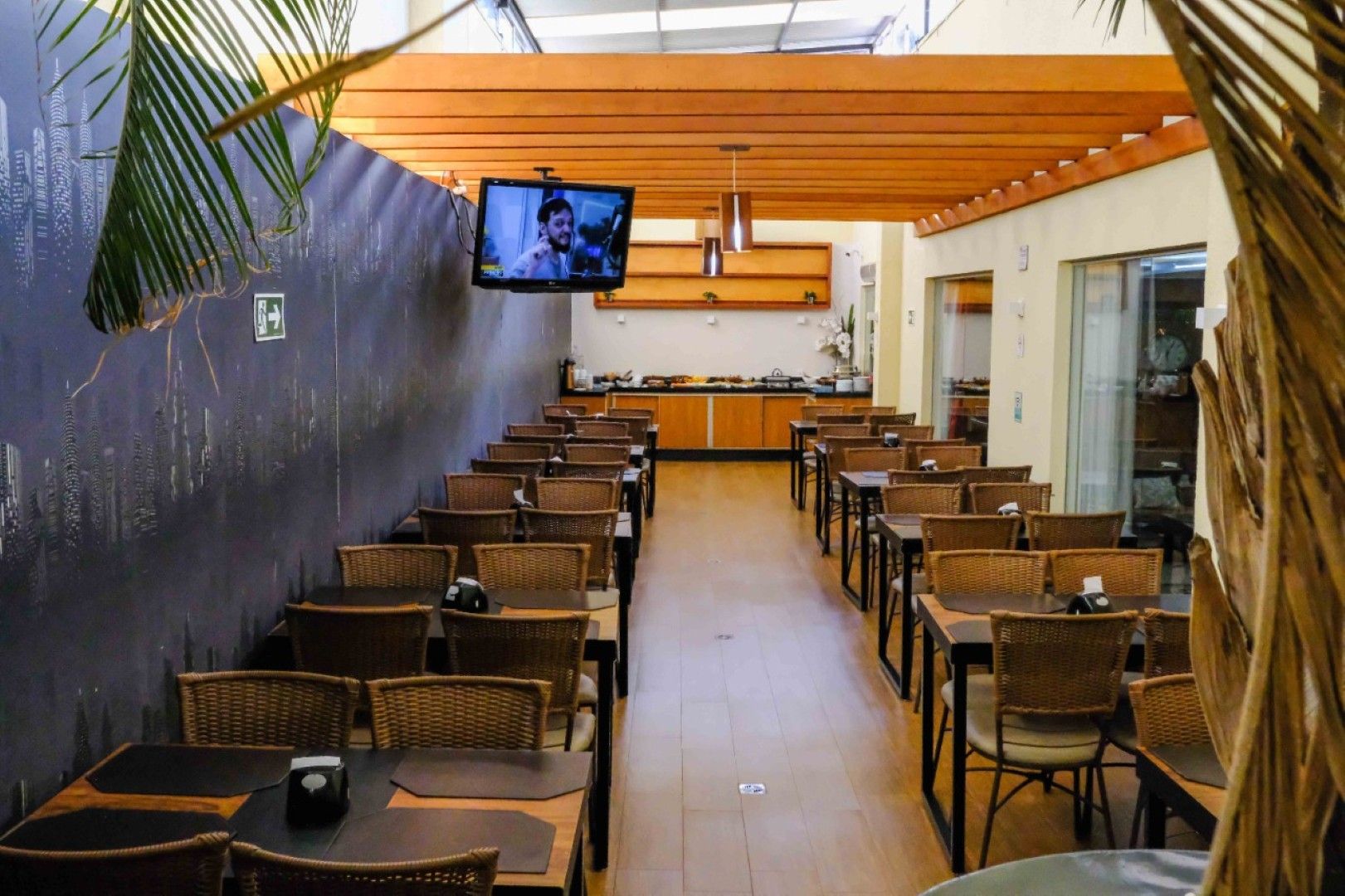 Café vazio com cadeiras de vime, mesas de madeira, TV de parede e balcão em uma área de refeições interna bem iluminada.