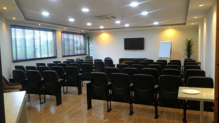 Sala de conferências vazia com fileiras de cadeiras pretas, tela fixada na parede, quadro branco e iluminação aconchegante.