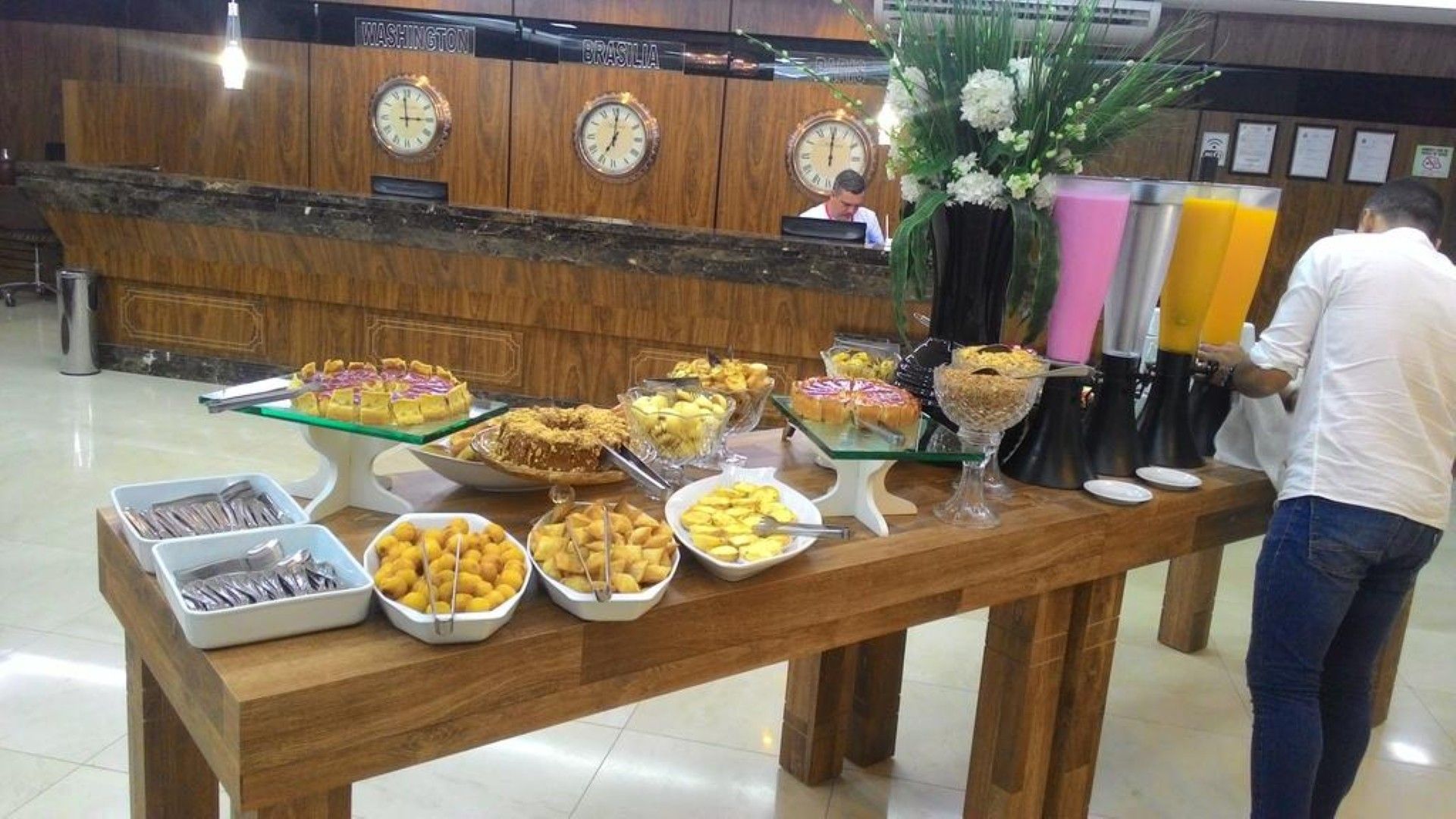Mesa de buffet com bandejas de comida variada e frutas, ao lado de dispensadores de bebidas rosa e pretos em um hall.