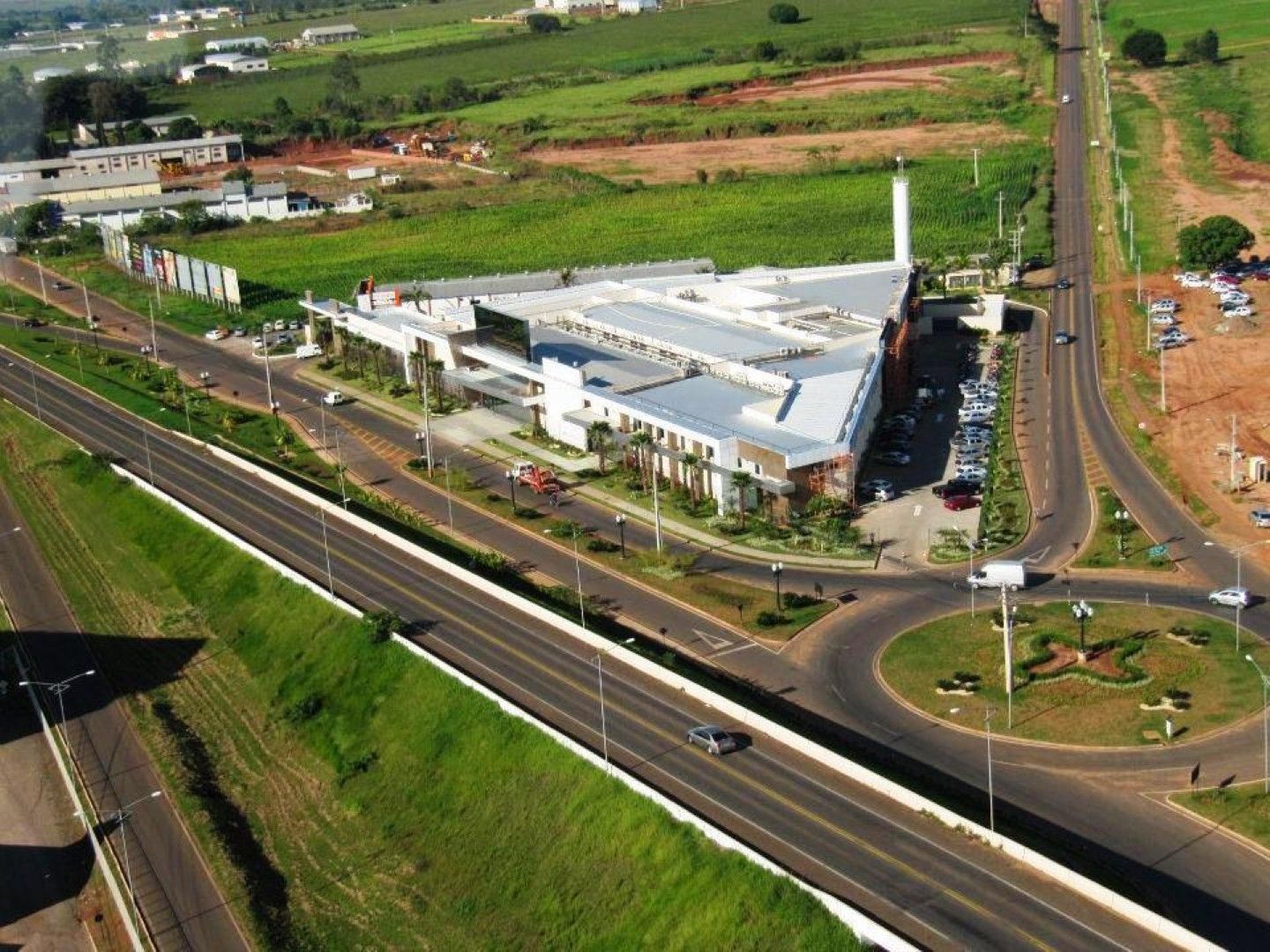 Vista aérea de um edifício branco ao lado de uma rodovia e rotatória em uma área rural verde.