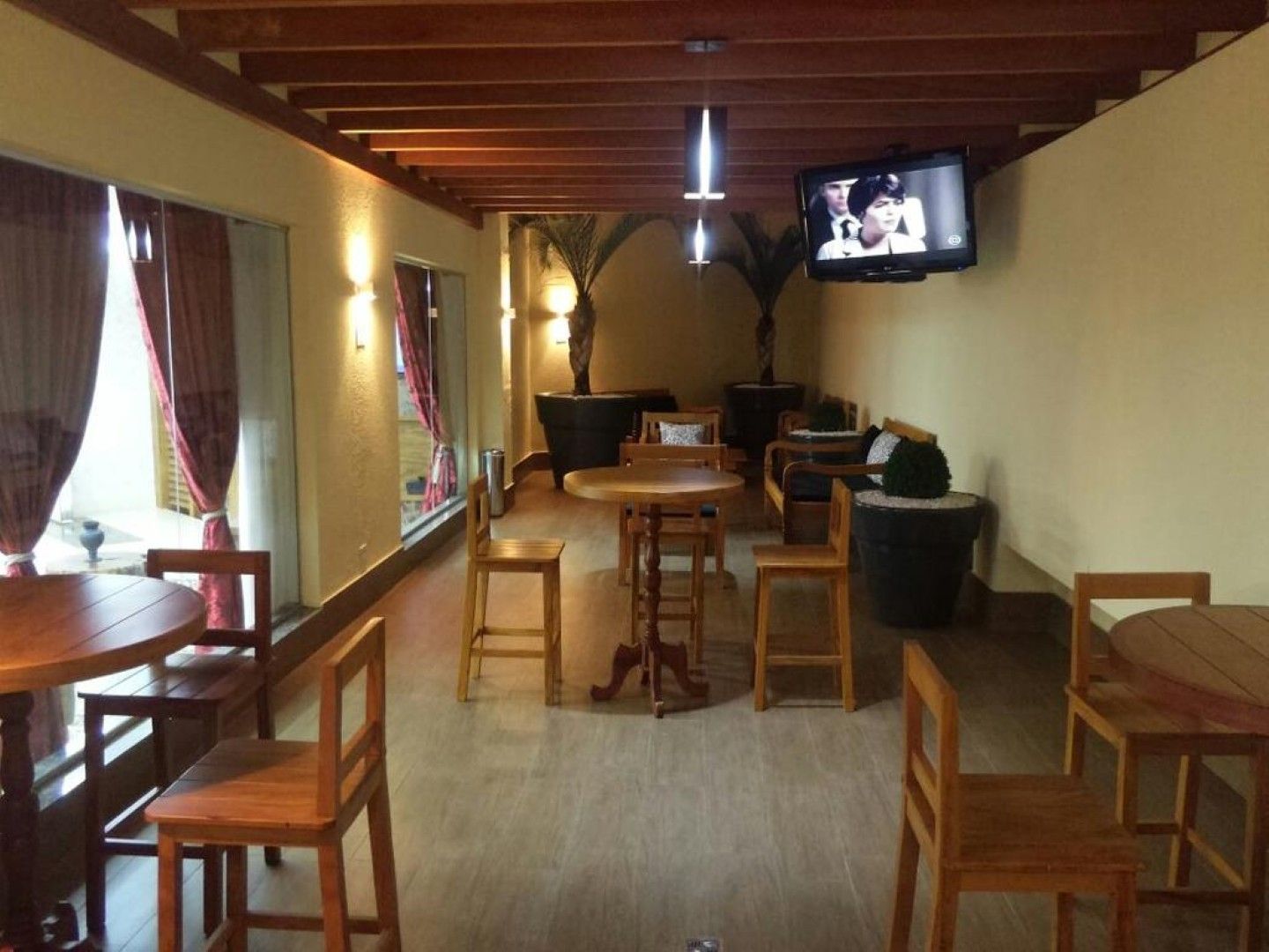 Interior aconchegante de um café com mesas e cadeiras de madeira, cortinas e uma TV fixada na parede acima.