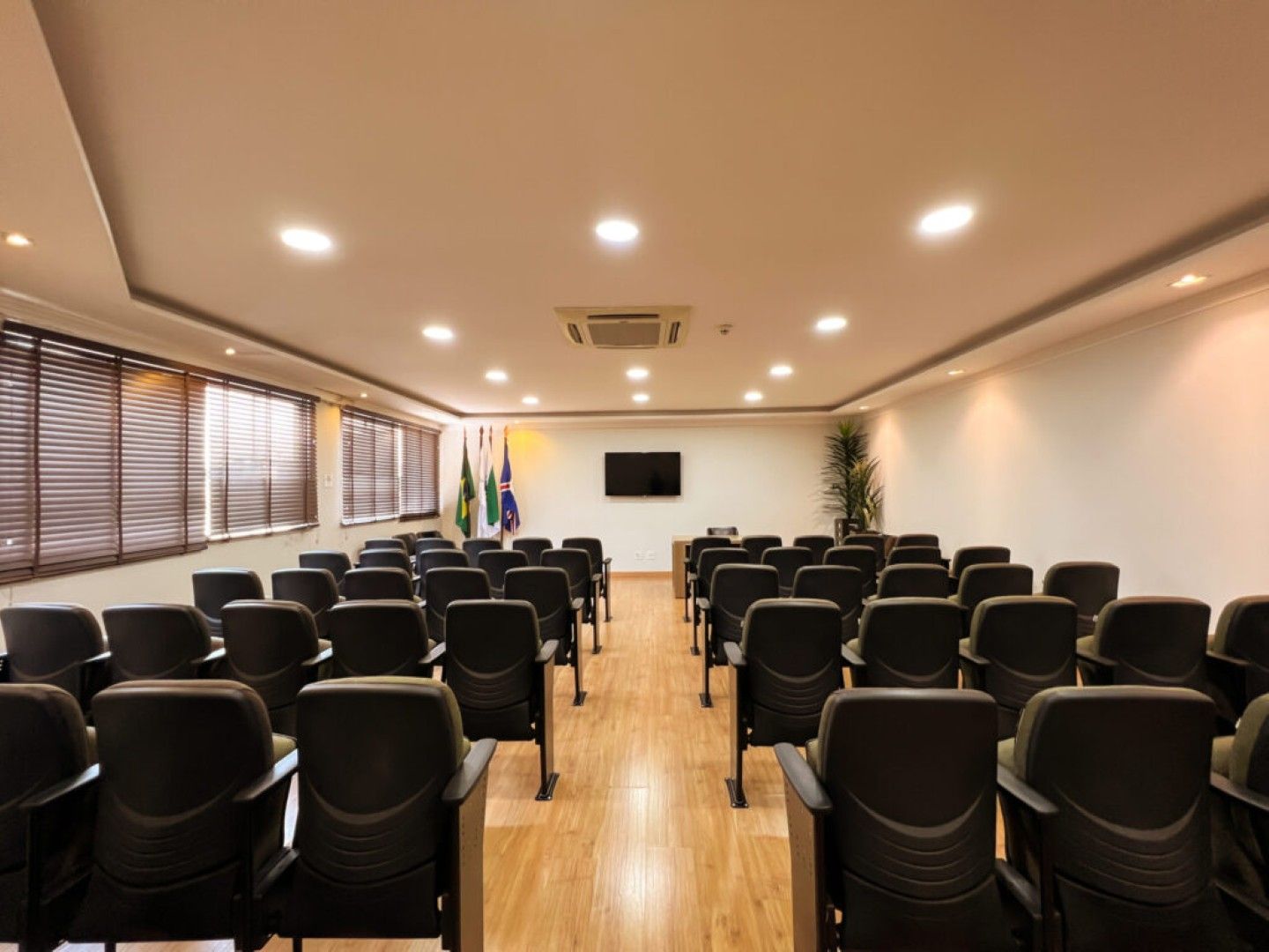 Sala de conferências vazia com fileiras de cadeiras pretas, piso de madeira, luzes no teto e uma tela fixada na parede.