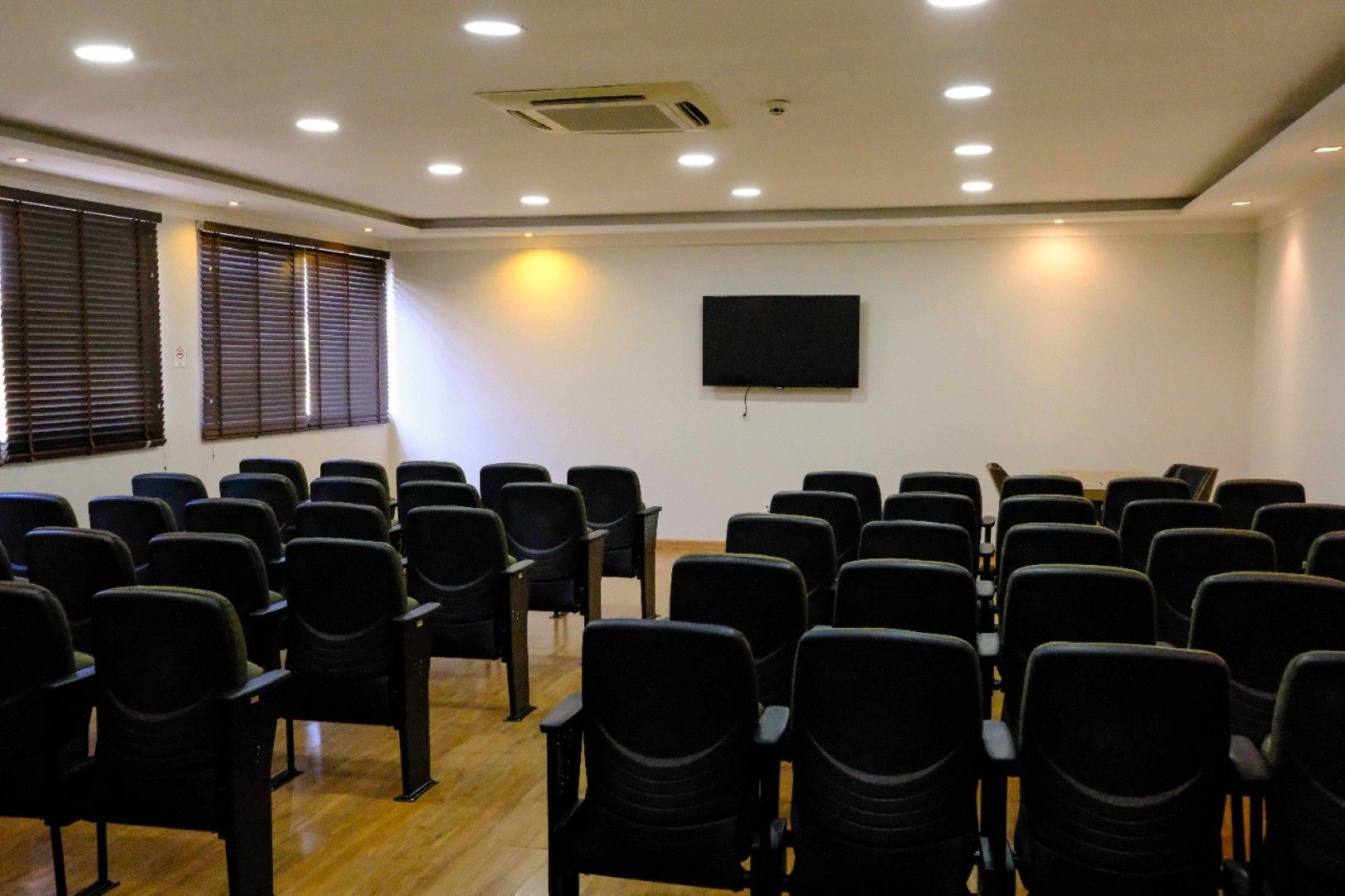 Sala de conferências vazia com fileiras de cadeiras pretas, TV fixada na parede e persianas.