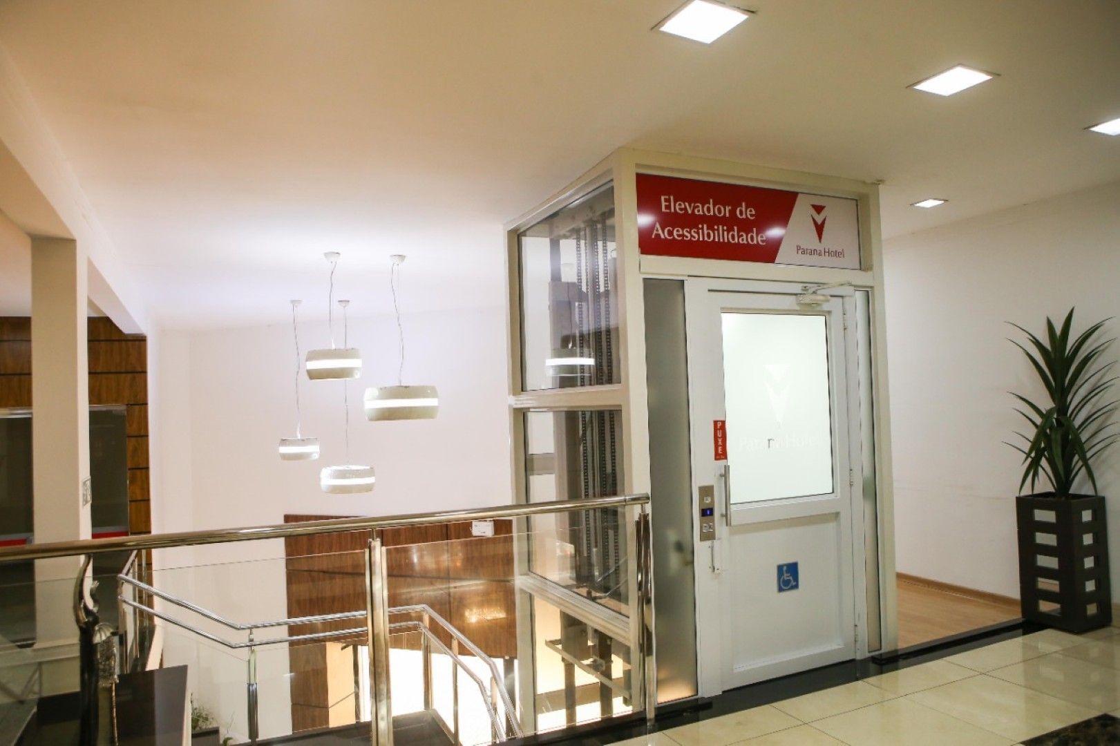 Átrio interno iluminado com elevador de vidro, corrimão de escada e vaso de planta ao lado da porta.