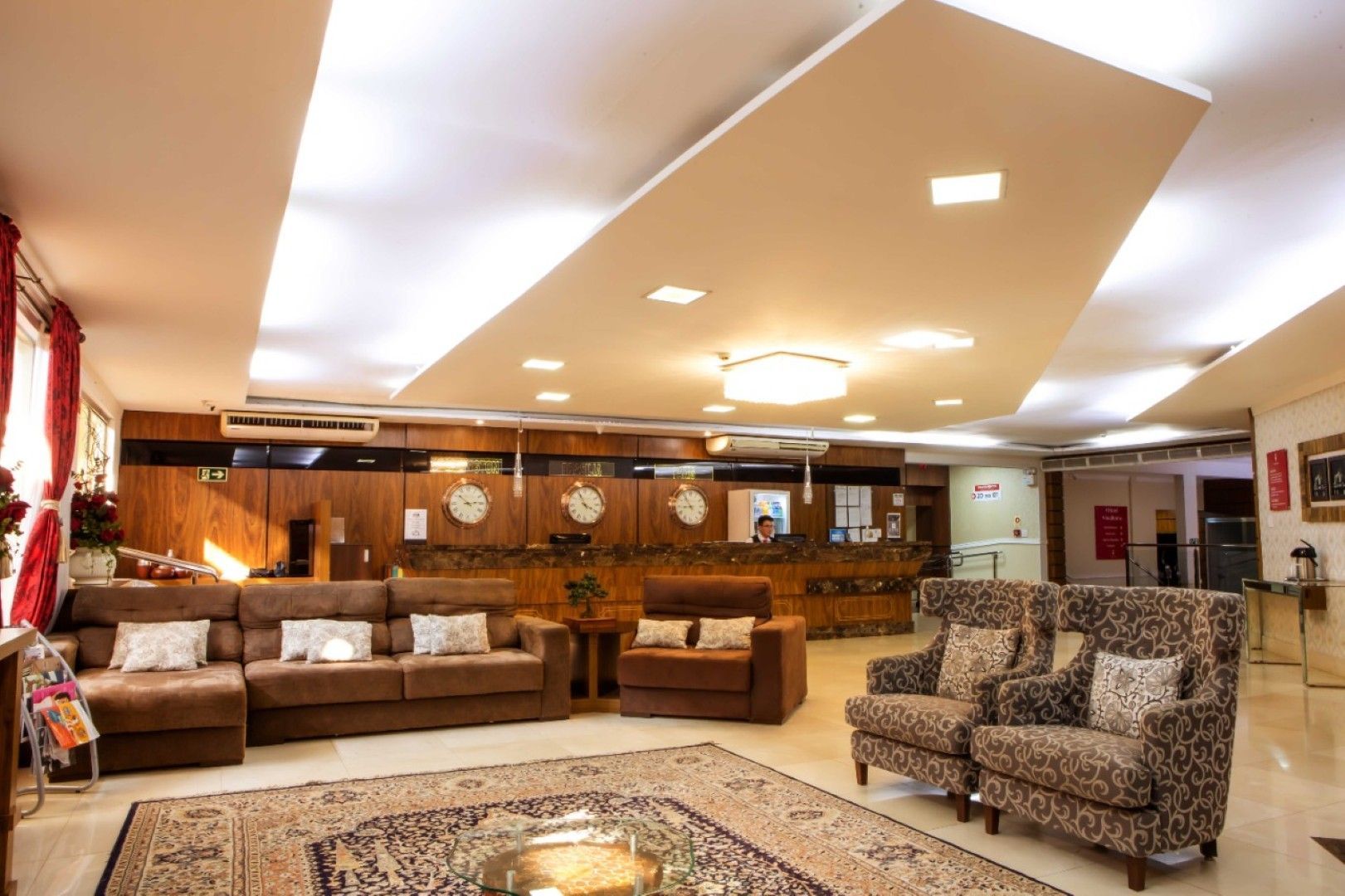 Amplo lobby do hotel com sofás bege, cadeiras estampadas e balcão de recepção com painéis de madeira sob luzes de teto aconchegantes.