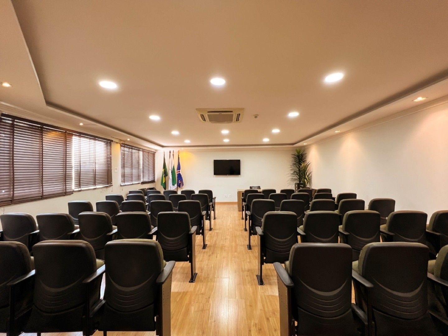 Sala de conferências vazia com fileiras de cadeiras pretas, piso de madeira, TV fixada na parede e bandeiras perto da entrada.