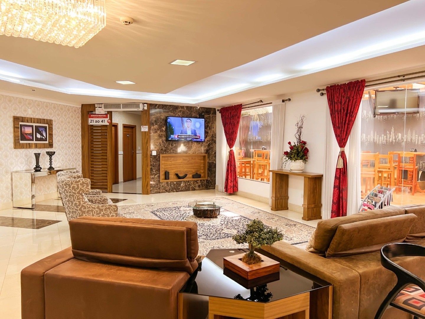 Elegante hall de entrada do hotel com poltronas de couro bege, lareira, TV e cortinas vermelhas com vista para a cidade.
